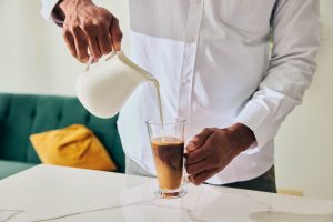 Man pouring milk into a glass of warm drink at a kitchen counter
